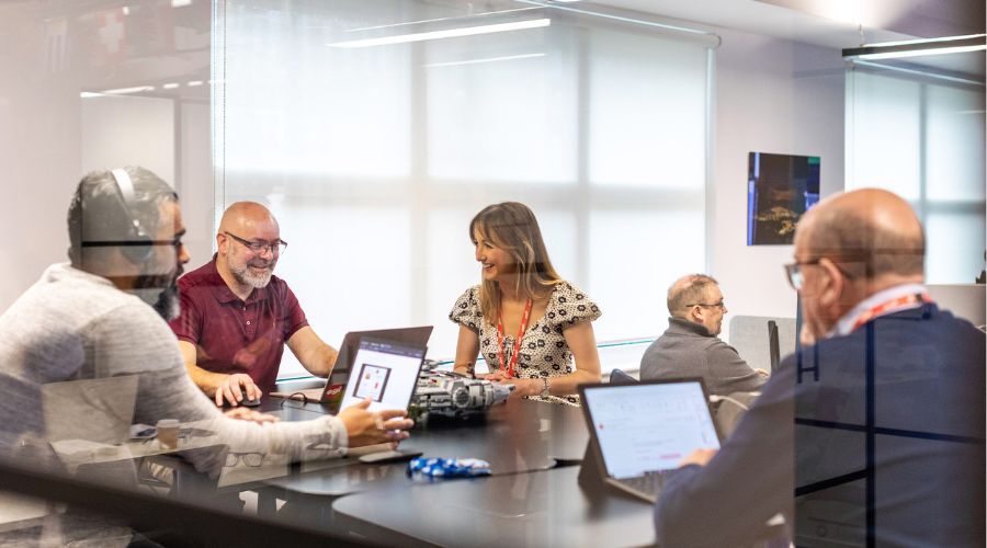 Several employees working at a desk using modern workplace solutions, like laptops and headsets, to collaborate together. 