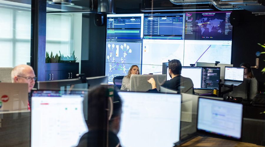 Employees working at desktops amidst several large screens, using modern workplace solutions in the office. 