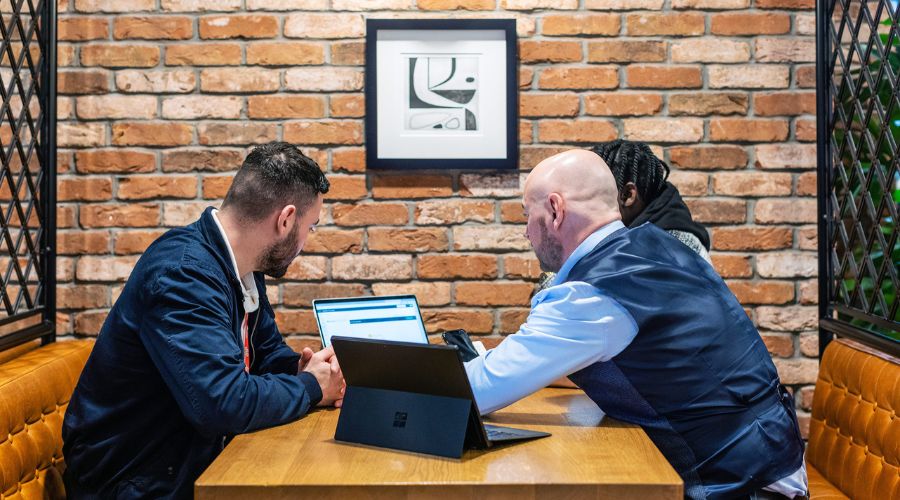 Three employees at a desk circled around two laptops, using modern workplace solutions to improve their productivity levels.