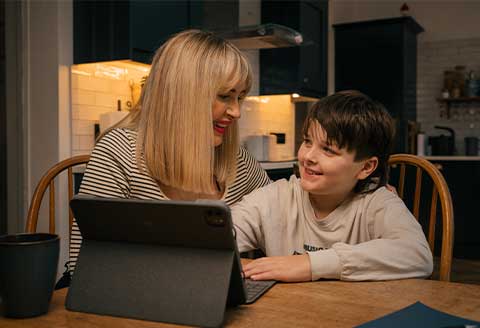 A mother an son at the kitchen table in front of an iPad. They are looking at each other smiling. Behind them a dimly lit modern kitchen can be seen.)